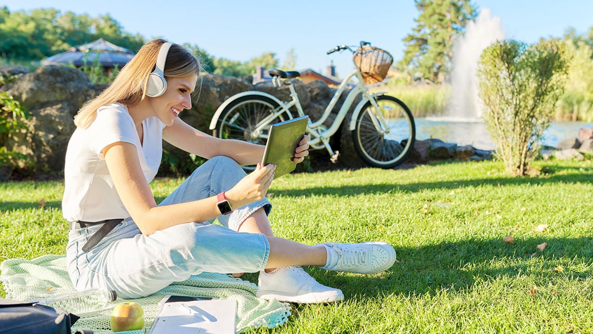 young woman in headphones with digital tablet looking in screen of gadget, video, film, call young woman in headphones with digital tablet looking in screen of gadget, video, film, call. Female sitting on green lawn in park on sunny summer day